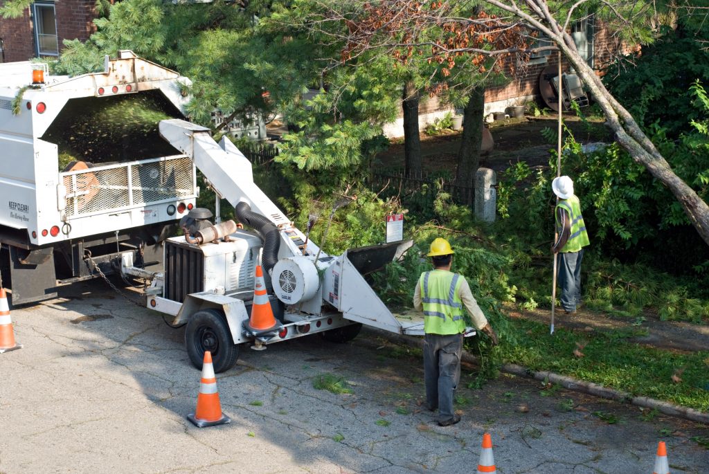 Two workers in safety gear operate a wood chipper near a 'Recycling Collection' truck in Suffolk County, NY, with orange cones marking the tree maintenance zone.