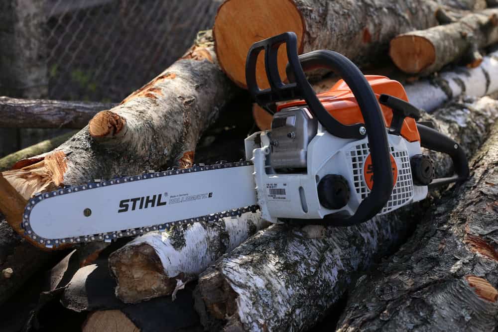 A chainsaw with an orange and white body rests on a pile of cut logs, perfect for tree trimming. The STIHL-branded tool stands out against the logs, which vary in color from gray to light brown. A chain link fence is visible in the blurred background, hinting at a neatly maintained landscape.